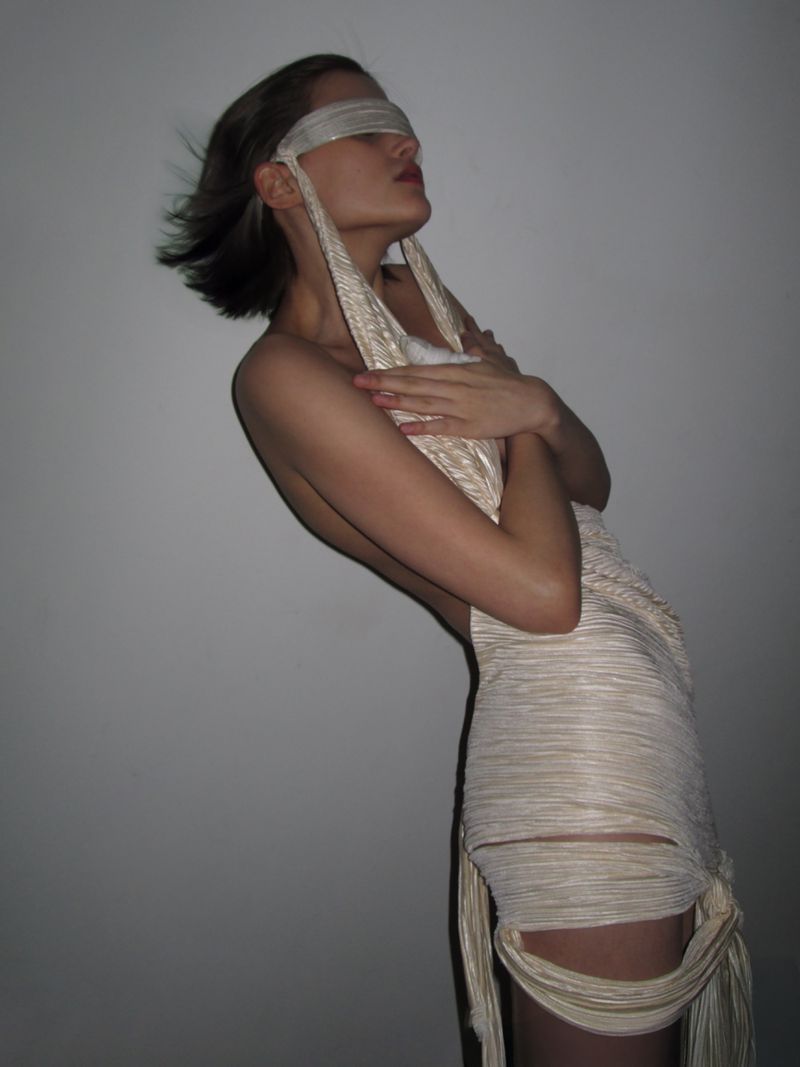 A woman with a blindfold poses in a light-colored, textured dress against a light gray backdrop, creating a minimalistic and evocative fashion image.