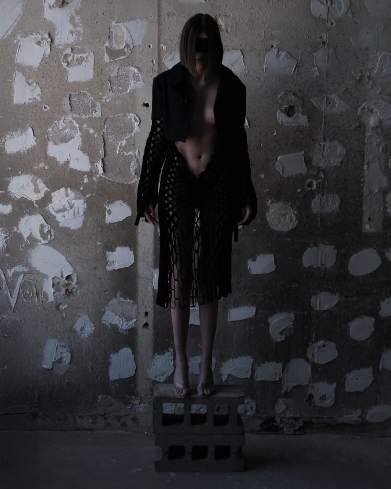 Barefoot woman in a black jacket and skirt poses on concrete blocks in a dilapidated interior with a textured and peeling wall.