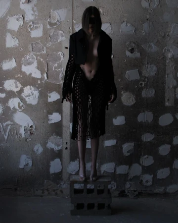 Barefoot woman in a black jacket and skirt poses on concrete blocks in a dilapidated interior with a textured and peeling wall.