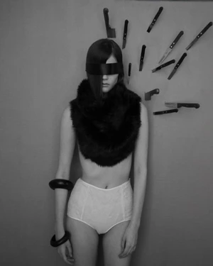 Blindfolded model wearing a black fur collar and high-waisted white briefs against a wall surrounded by embedded knives.