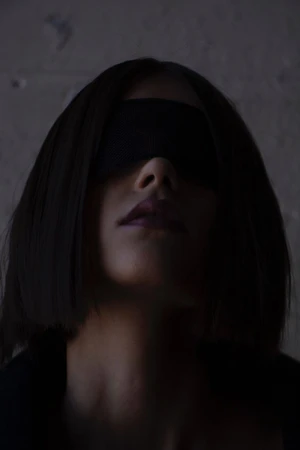 Close-up portrait of a woman with short dark hair wearing a black blindfold over her eyes against a dark textured wall