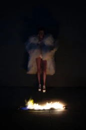 A model in an oversized white furry coat and platform heels standing in the dark behind a line of flames burning on the pavement.