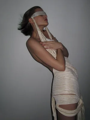 Model wearing wrapped cream textured dress with blindfold against a grey background for Coeval Magazine