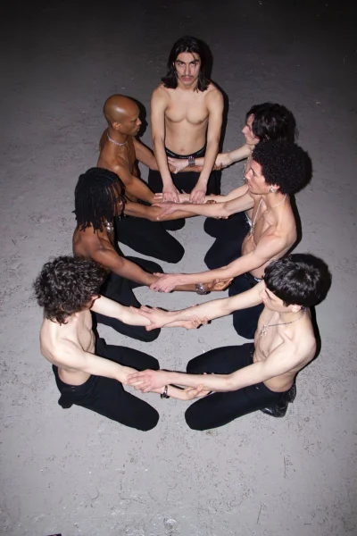 Seven shirtless young men in black trousers kneel in a circle holding forearms against a grey floor under direct flash lighting.