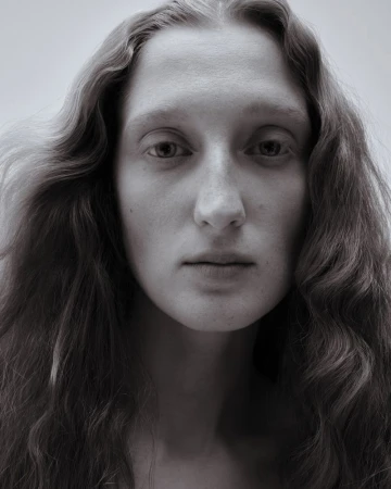 Close-up portrait of a woman with long wavy brown hair looking directly forward against a soft grey background.