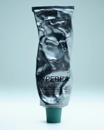 Crumpled metallic silver Typebea hair treatment tube standing upside down on a teal green cap against a pale blue background.