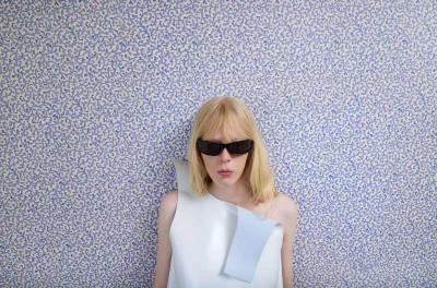 Blonde model wearing black narrow sunglasses and a sculptural asymmetrical white top against blue patterned wallpaper.