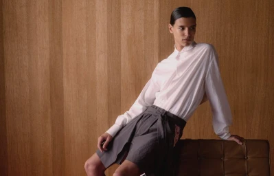Model Skander Gueye wearing a white collared shirt and grey pleated shorts leaning against a brown leather chair before a wood wall.