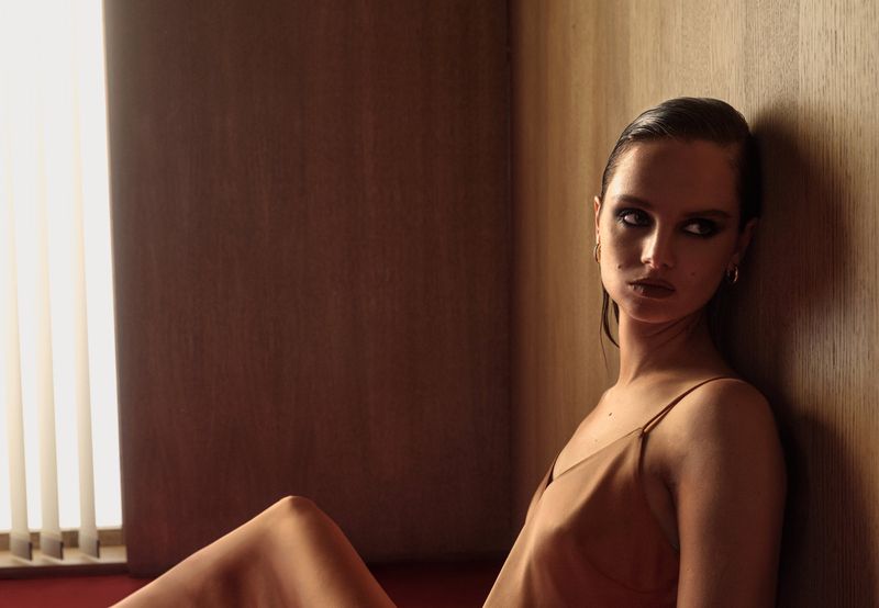 Model Giselle Norman leaning against a wood-paneled wall in a tan slip dress with smokey eye makeup under warm side lighting.
