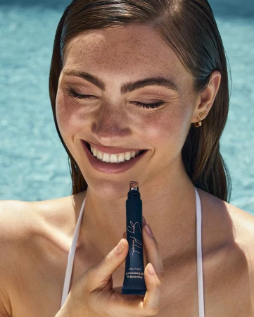 Smiling woman with wet hair and freckles holding a dark lip treatment tube in front of a blue swimming pool.