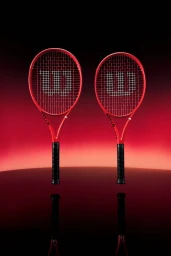 Two red Wilson tennis racquets standing upright side by side on a reflective surface against a dark red gradient studio background.