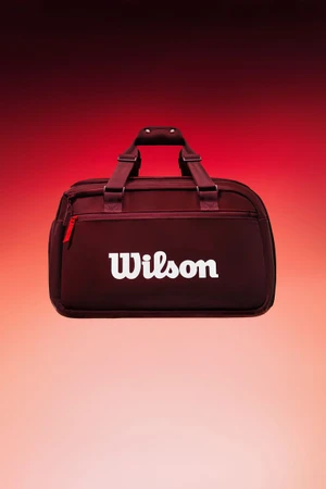 A burgundy Wilson duffel bag with a white logo centered against a gradient red and pink studio background.