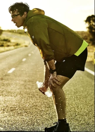 Man in an olive green hooded running jacket and black shorts resting hands on knees on an open asphalt road with a water bottle.