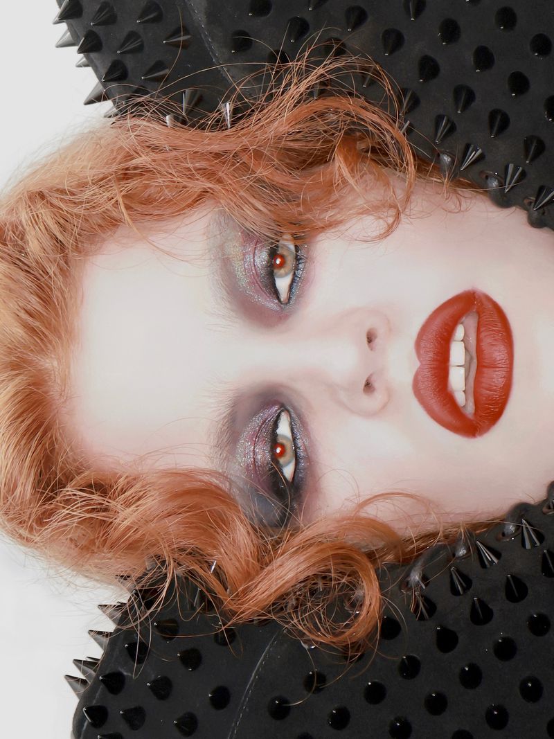 Close-up portrait of a woman with red hair and bold red lips, framed by black fabric decorated with numerous sharp, pointed spikes.