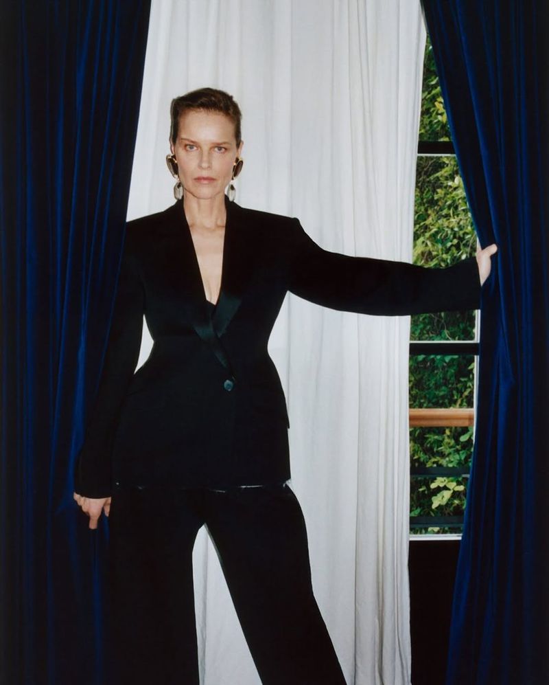 A woman in a black pantsuit stands between blue and white curtains against a green outdoor background, looking directly at the camera.
