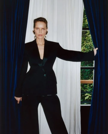 A woman in a black pantsuit stands between blue and white curtains against a green outdoor background, looking directly at the camera.