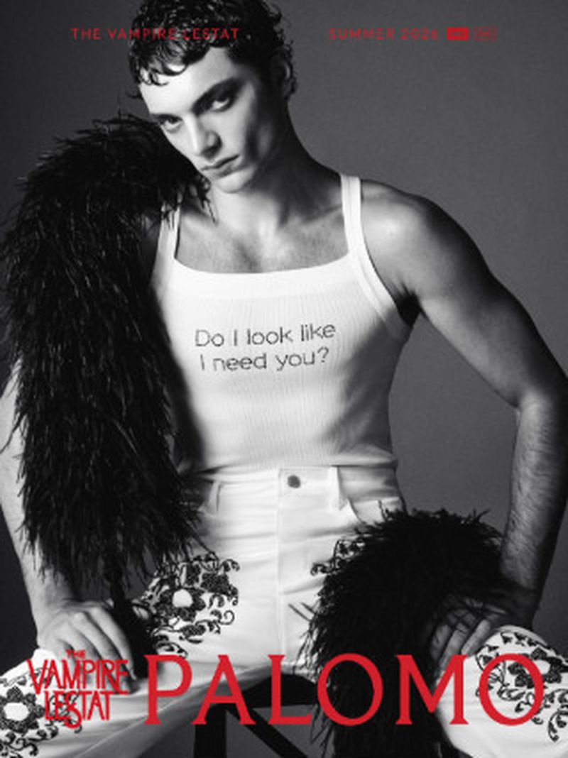 Male model in a white graphic tank top, embroidered trousers, and a black feather boa against a grey studio backdrop.