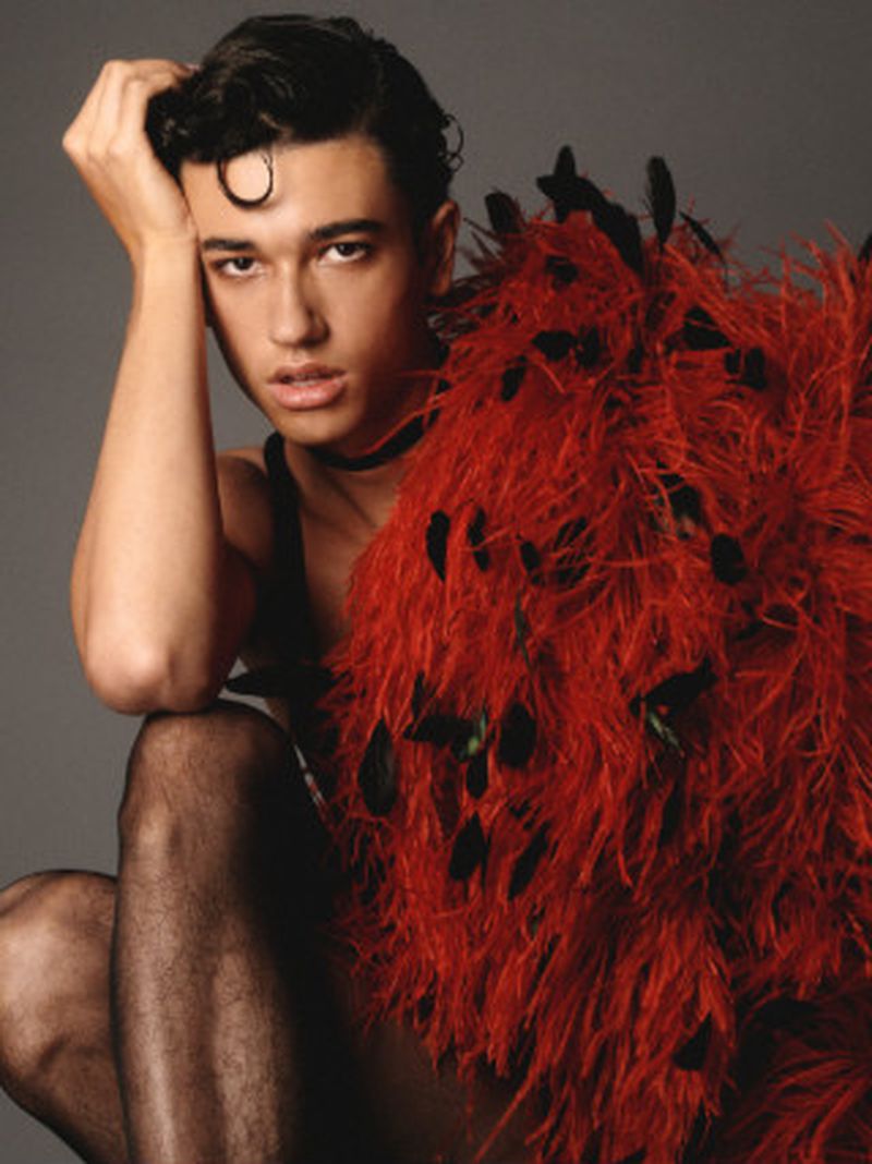 A young male model with dark hair and a curled fringe wears a red feathered shawl and a black choker against a grey backdrop.