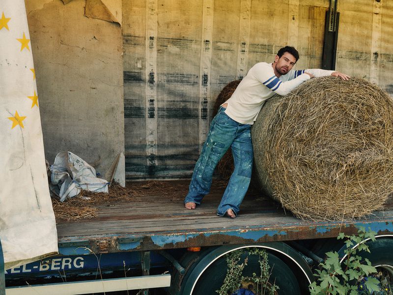 Theo James leaning against a large round hay bale on the back of a truck, wearing a white long-sleeve shirt and distressed denim jeans.