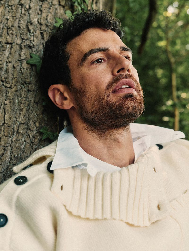 Close-up of Theo James leaning against a tree, wearing a cream-colored ribbed knit sweater with dark buttons over a white shirt.