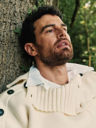 Close-up of Theo James leaning against a tree, wearing a cream-colored ribbed knit sweater with dark buttons over a white shirt.