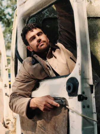 Theo James wearing a beige jacket, leaning out of a white van door with a neutral expression.