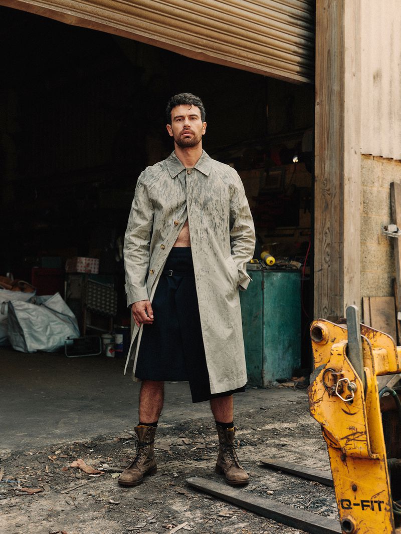 Theo James wears a paint-splattered trench coat and navy kilt standing in front of a rustic garage with a yellow forklift blade.