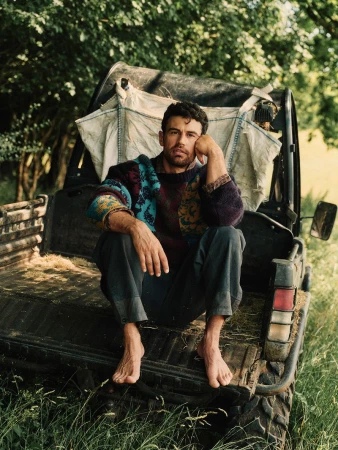 Theo James sitting barefoot in the back of a utility vehicle wearing a patterned knit sweater and trousers in a rural outdoor setting