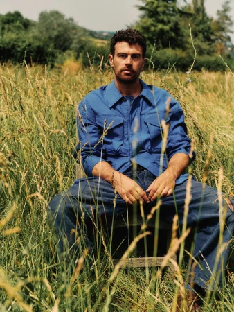 Theo James wearing a blue button-down shirt and trousers seated on a wooden chair in a tall, sunlit meadow of green and yellow grass.