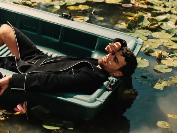 Theo James lying in a green plastic boat, wearing a black robe with white piping, surrounded by lily pads on calm water.