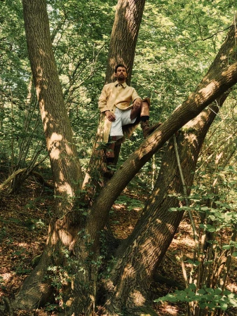 Theo James wearing a tan buttoned jacket and white shorts perched in the fork of a large forest tree for British GQ.
