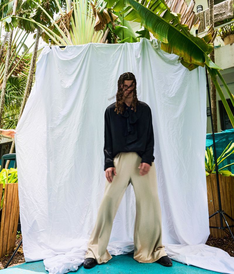 A person with braided hair wears a black silk shirt and wide-leg trousers standing before a white backdrop in a garden setting