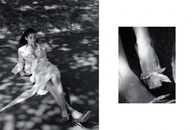 Model Raynara Negrine reclines outdoors in a satin wrap coat dress with bow-accented heels in a black-and-white editorial spread.