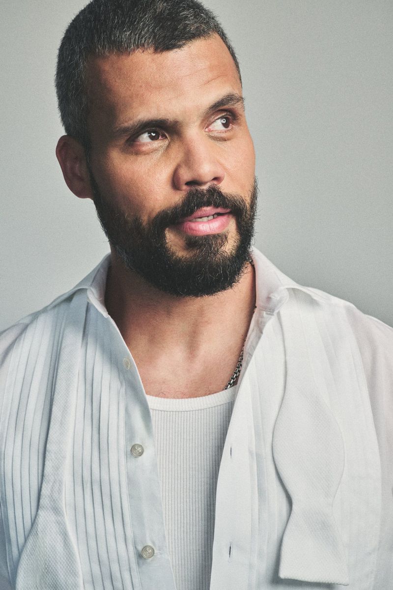 Howard Charles wearing an unbuttoned white pleated tuxedo shirt with an untied bow tie against a grey backdrop.