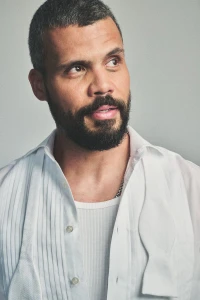 Actor Howard Charles in an unbuttoned white pleated shirt and untied bow tie against a grey studio backdrop.
