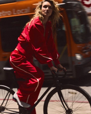 A woman with wavy hair wearing a vibrant red tracksuit rides a black bicycle on a sunlit urban street in front of an orange city bus.