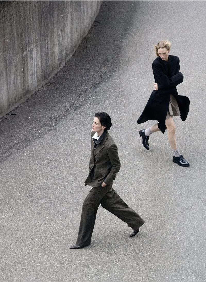 Two fashion models in dark tailored suits and coats walking across an asphalt road next to a concrete wall.