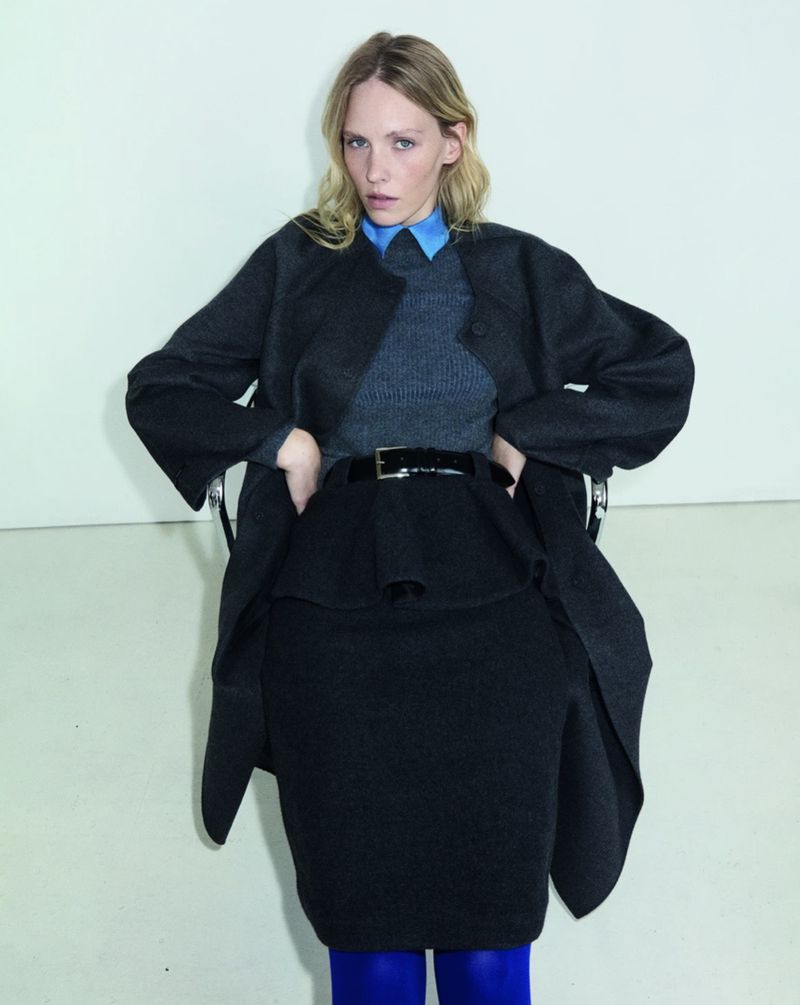 A blonde woman sitting on a chair, wearing a charcoal wool coat over a grey sweater, blue collar shirt, and black skirt against a plain wall.