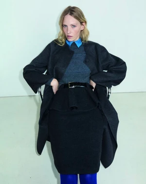 Model seated in a charcoal wool coat and peplum skirt with a blue collared shirt and vibrant cobalt tights against a pale backdrop.