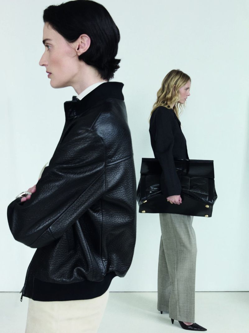 Two fashion models standing in profile wearing tailored black garments and an oversized black bag against a white studio background.