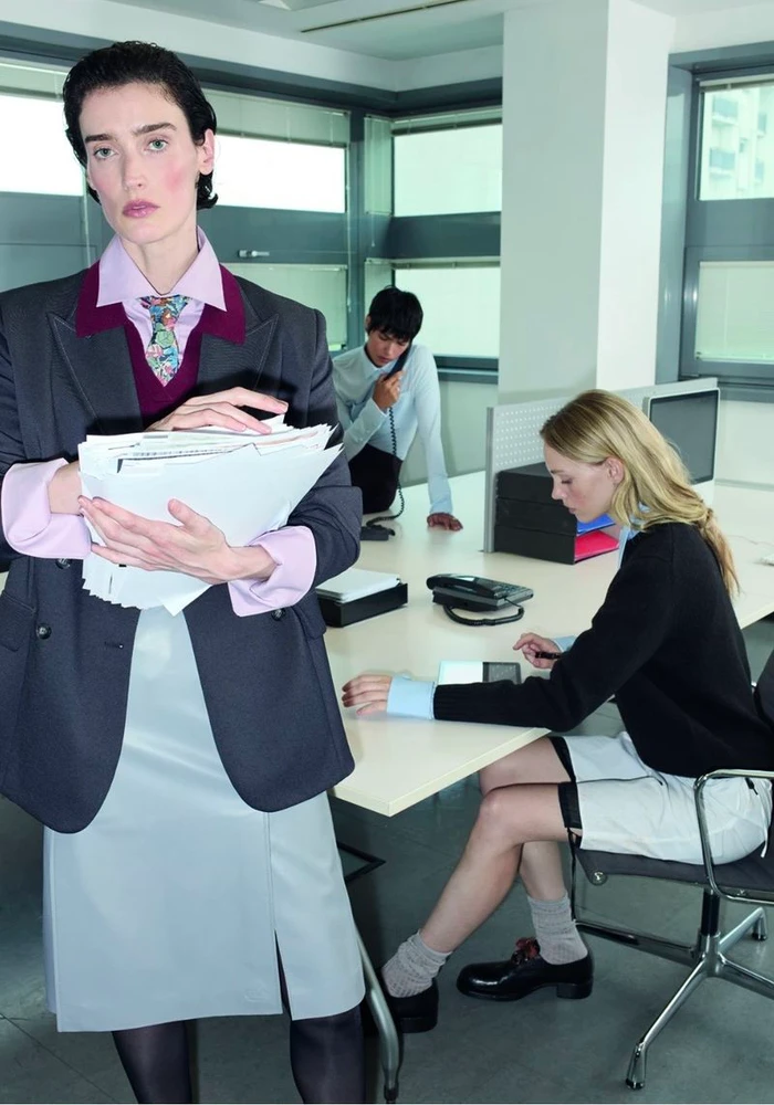 Model holding a stack of papers in an office setting wearing a charcoal blazer, pink shirt, tie, and light grey skirt.