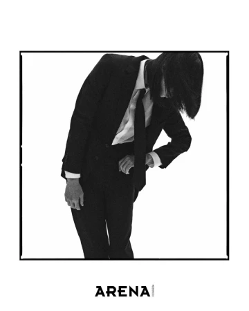 A person in a black suit with long hair leans forward in a white background, captured in a stylish black-and-white studio portrait.