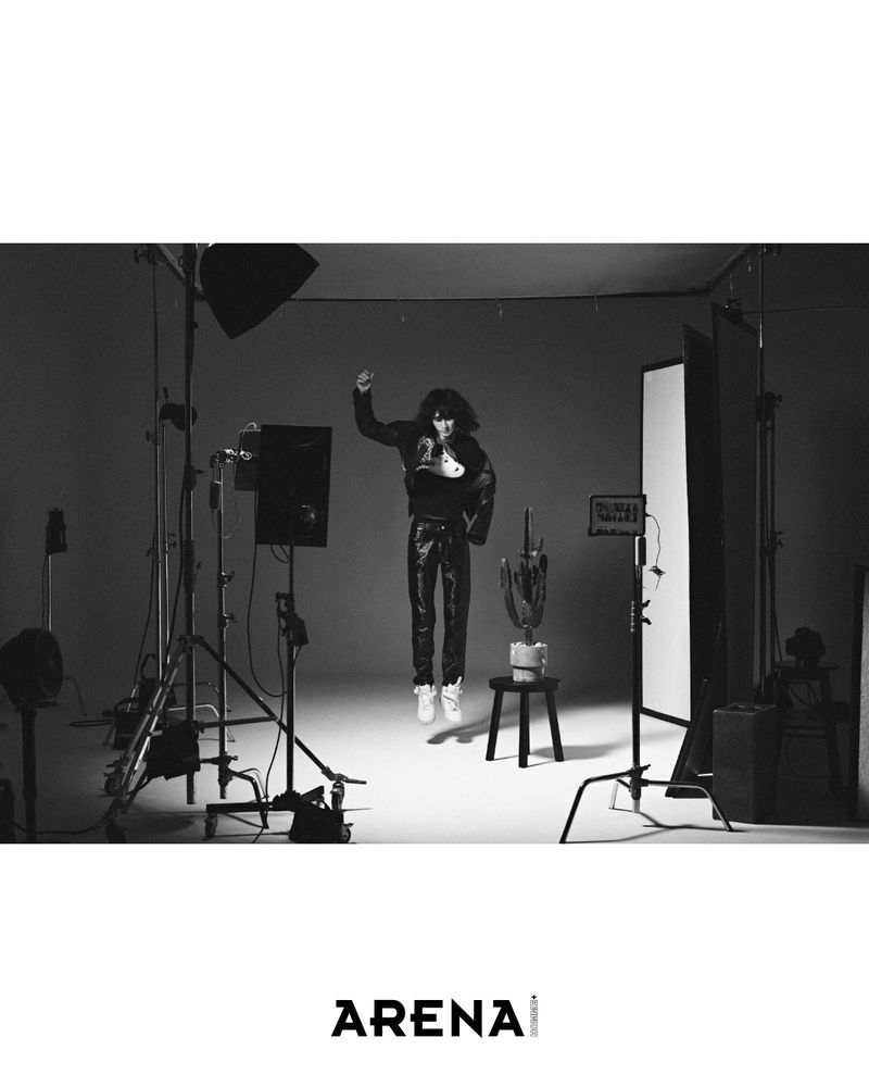 Black and white photo of person mid-air in black leather-like pants and jacket in a photo studio with lights and equipment.