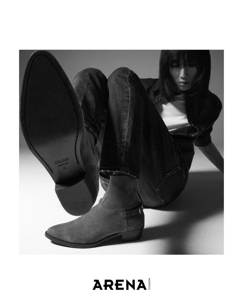 Black and white fashion shot: woman poses with Celine ankle boots, blue denim jeans, white top, and a dark jacket indoors.