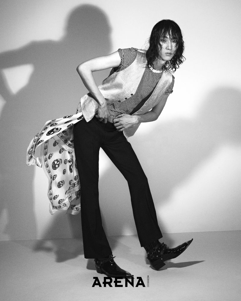 Model Han Ji poses in a vest, flared trousers, skull-print scarf, and pointed harness boots against a studio backdrop.