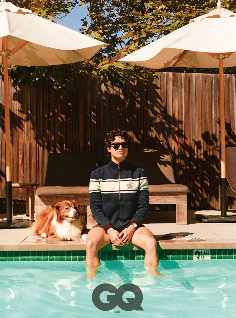 Charles Melton sitting at the edge of a swimming pool in sunglasses and a navy jacket beside a brown and white dog.