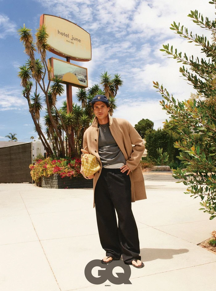 Charles Melton standing outdoors in a camel coat, black trousers, and a baseball cap near a Hotel June sign for GQ Korea.