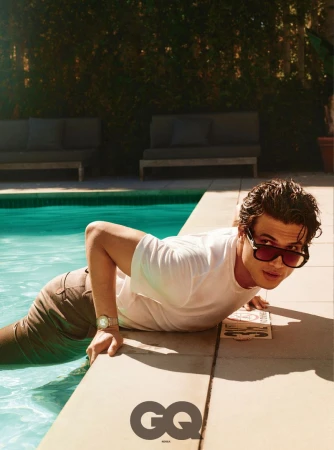 Actor Charles Melton wearing a white t-shirt and sunglasses while leaning on the edge of an outdoor swimming pool.