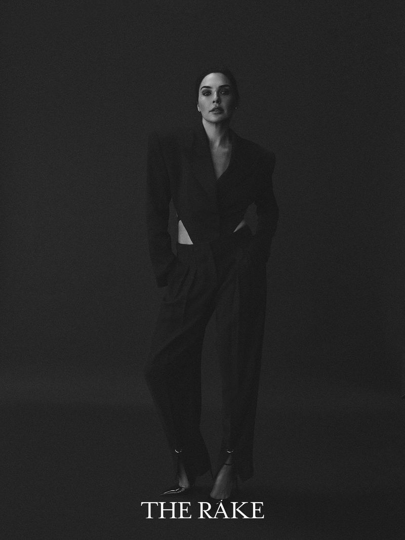 Gal Gadot posing in a black cutout blazer and tailored trousers against a dark background for The Rake.