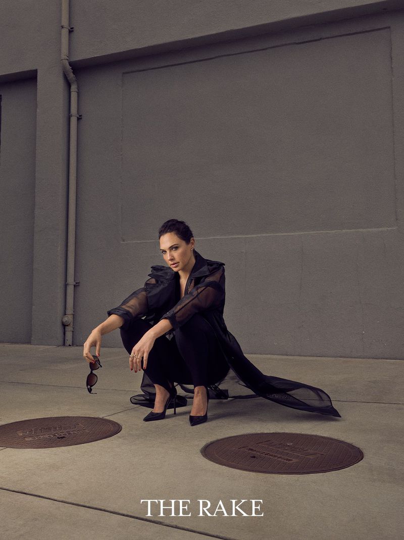 Gal Gadot crouching on concrete in a sheer black coat, black pants, and pointed heels holding sunglasses.